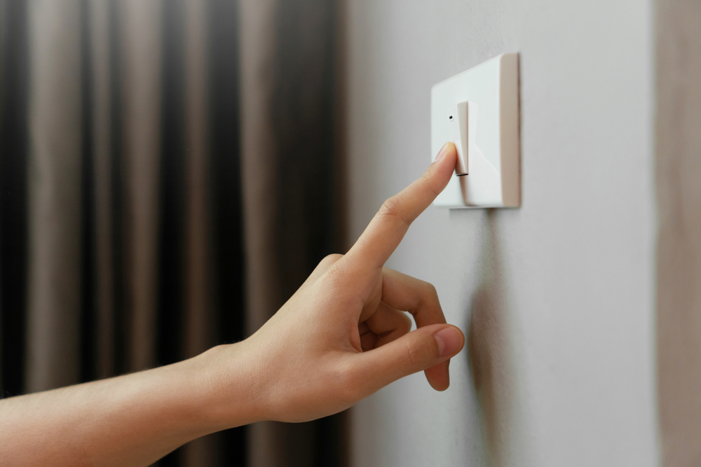 Casa a risparmio energetico: 5 tips. 1 Dettaglio di mano su interruttore luce; l'interruttore e la parete su cui è installato sono bianchi. Sullo sfondo si intravede una tenda di colore marrone.